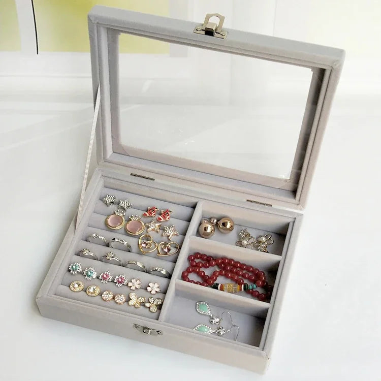 Light gray velvet jewelry organizer with clear lid containing rings, earrings, and a red beaded bracelet